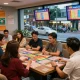 Penjudi yang bersemangat mendiskusikan hasil Pengeluaran HK Lotto di sebuah lounge yang ramai dengan tiket lotere dan layar digital.