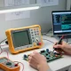 Engineer measuring signals with a handheld oscilloscope multimeter in a tech workshop.